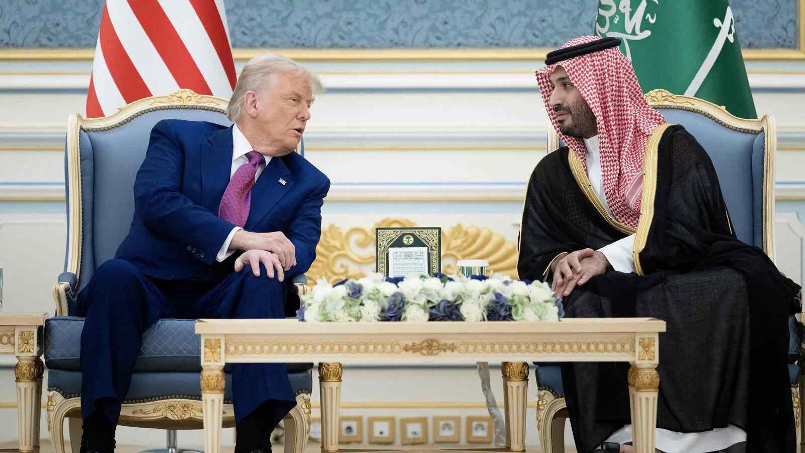 Former U.S. President Donald Trump and a Saudi official in traditional dress sit and talk in an ornate room with American and Saudi flags in the background, and a table with flower arrangements in front of them.